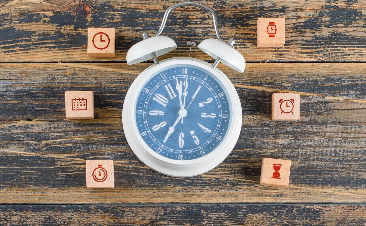 Time management concept with wooden blocks with icons, big clock on wooden table flat lay.