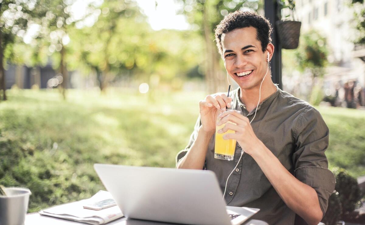 Cheerful guy with laptop and earphones sitting in park while drinking juice and smiling at camera