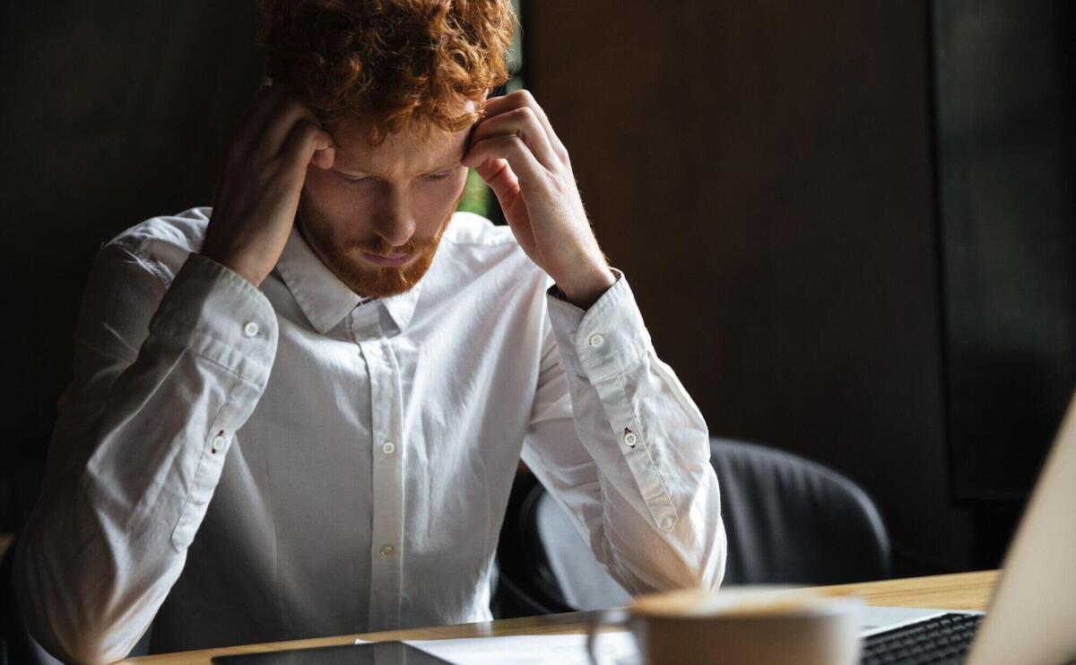 Photo of tired redhead businessman, touching his head, looking at digital tablet