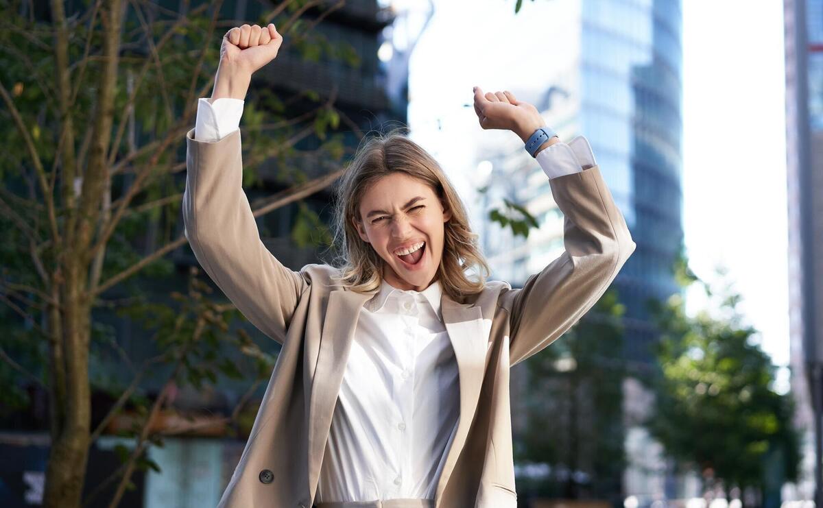 Excited businesswoman screams and celebrates lifts hands up does hooray gesture celebrates victory achieve goal stands on street