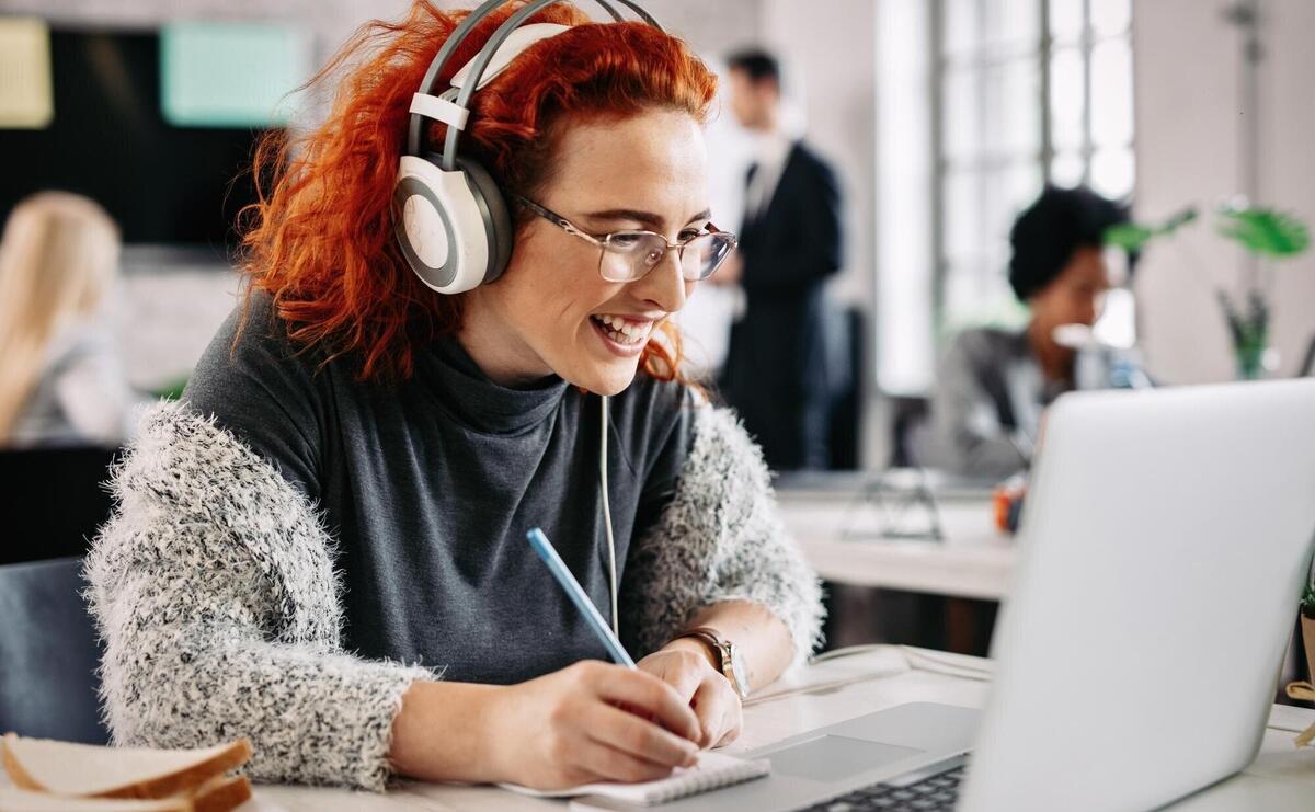 Cheerful female entrepreneur working on a computer and taking notes in her notepad while listening music on headphones in the office There are people in the background