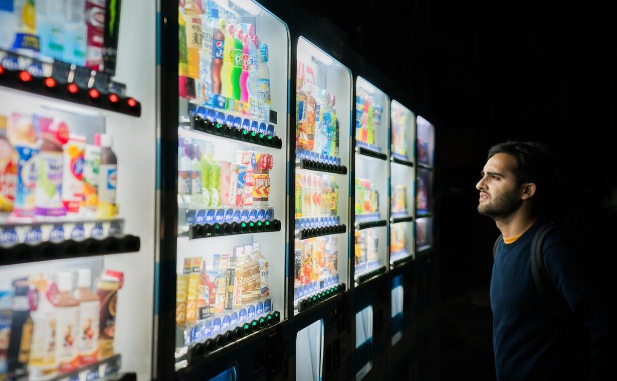 Man gazes at colorful vending machines, indecisive under the neon glow.