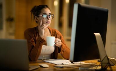 Young woman drinking coffee and thinking while working on desktop PC in the evening at home