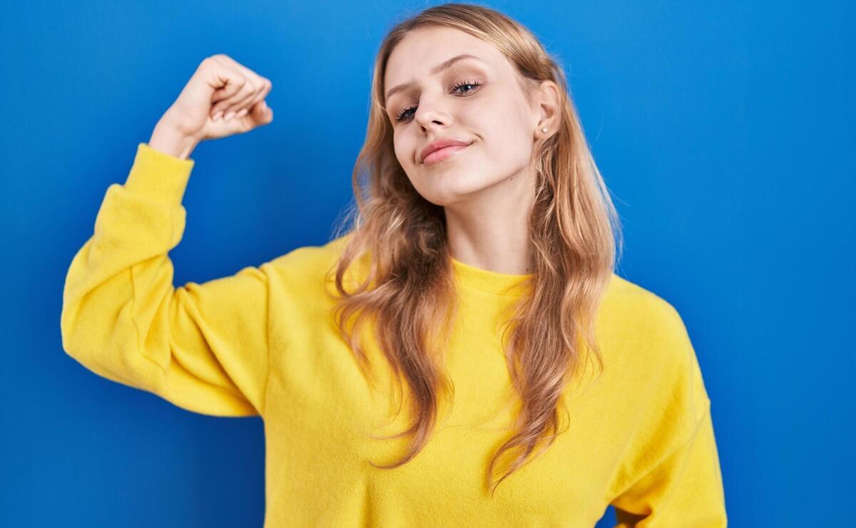 Young caucasian woman standing over blue background strong person showing arm muscle, confident and proud of power