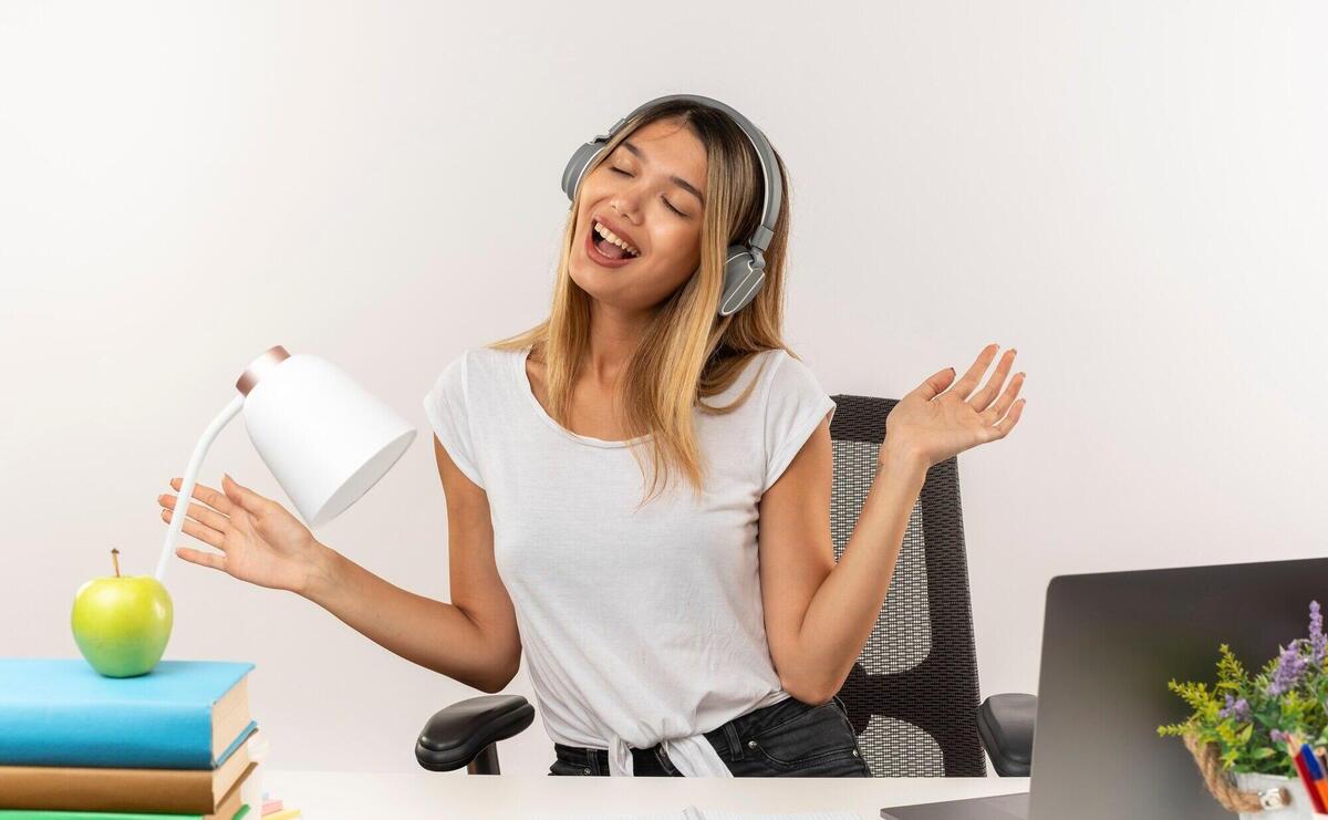 Joyful young pretty student girl wearing headphones sitting at desk with school tools listening to music showing empty hands with closed eyes isolated on white