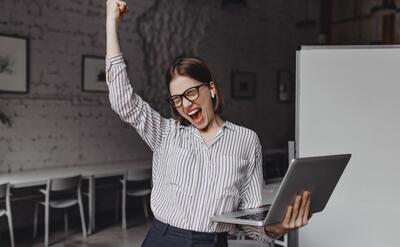 Business woman with laptop in hand is happy with success. Portrait of woman in glasses and striped blouse enthusiastically screaming and making winning gesture.