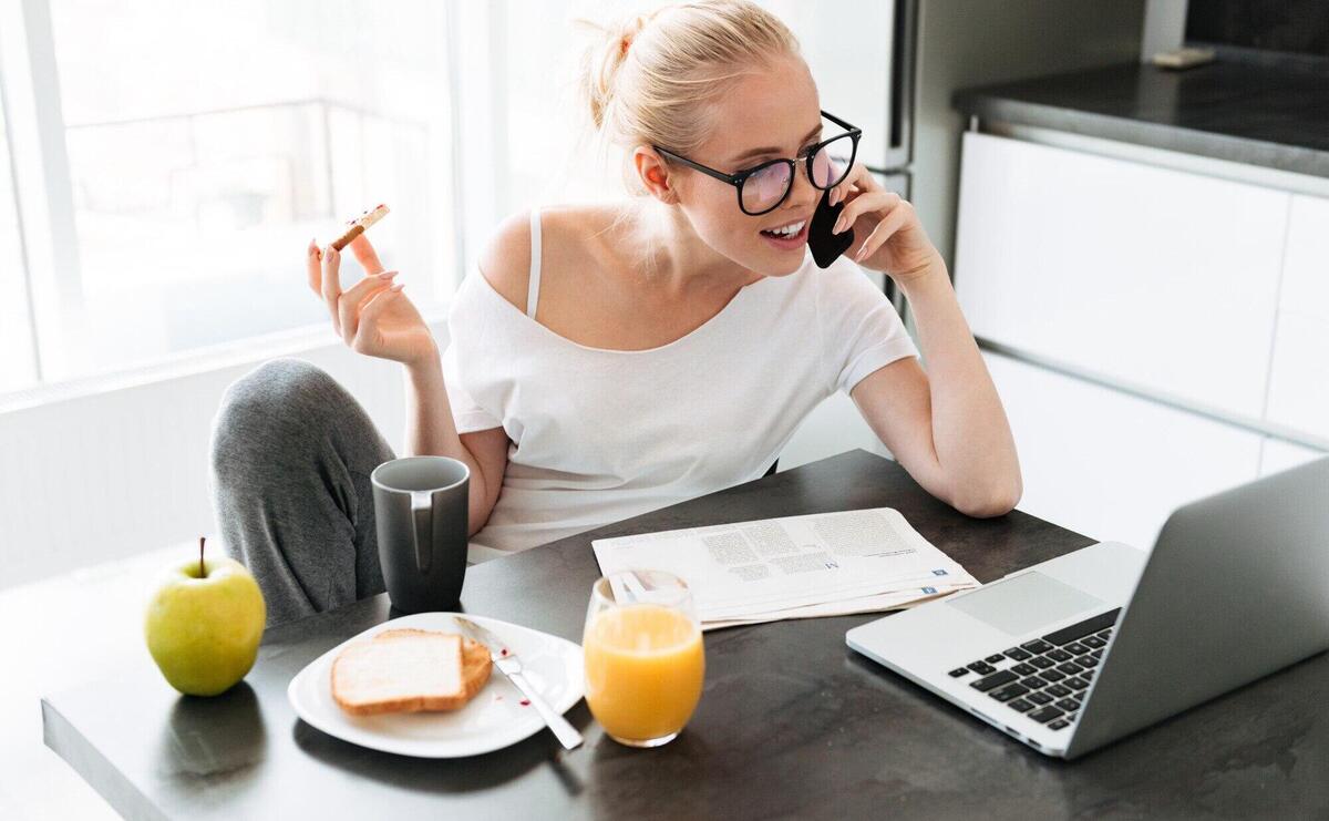 Beuatiful lady working with laptop and talking on smartphone and eating breakfast