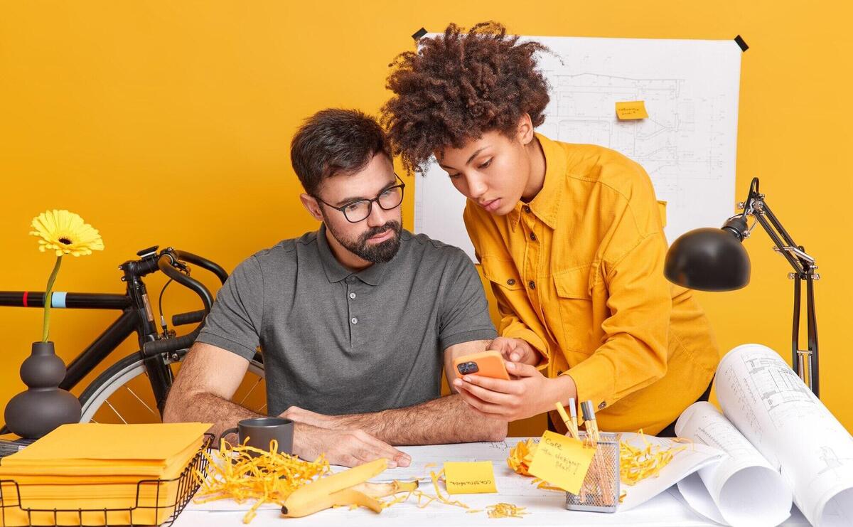 Two diverse members of working team think over cretive ideas for future project woman shows design plan in smartphone on webpage pose at desktop 