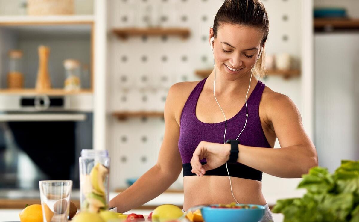 Happy athletic woman checking time on her wristwatch while preparing fruit smoothie in the kitchen