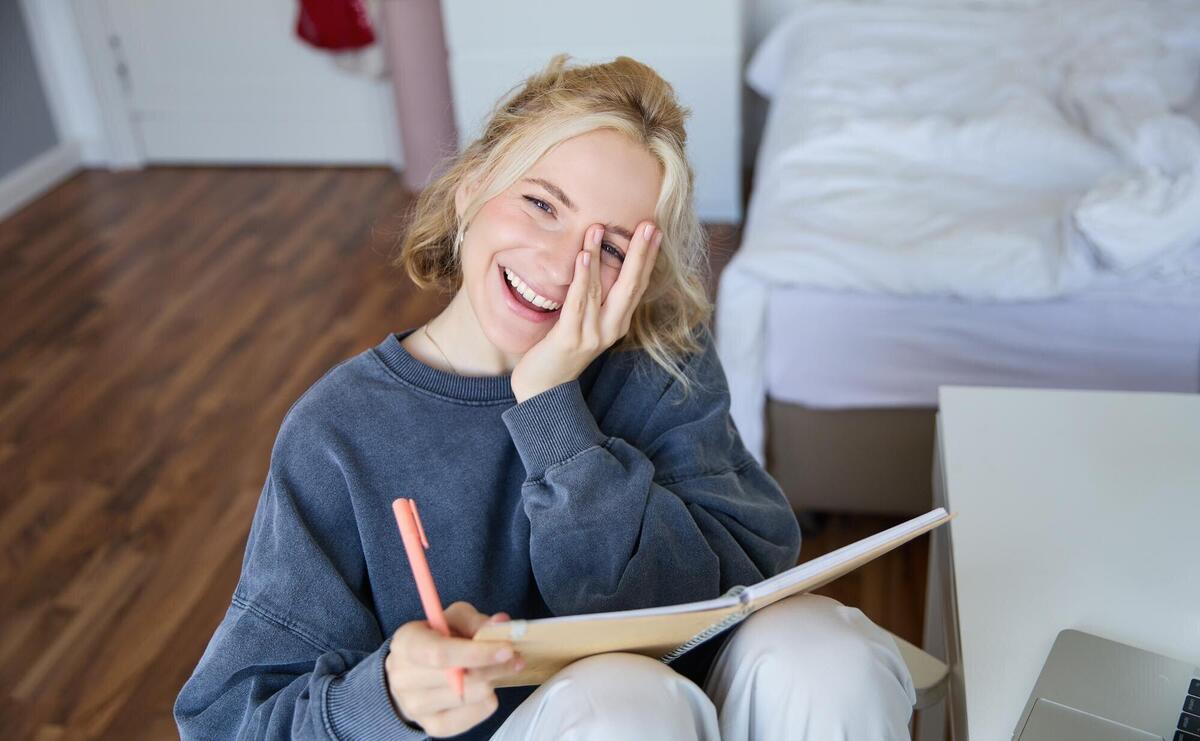 Portrait of happy young blond woman sitting in bedroom with notebook and pen laughing and smiling