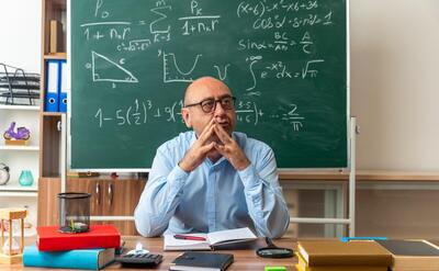 impressed looking side middle-aged male teacher wearing glasses sits at table with school supplies holding hands together in classroom