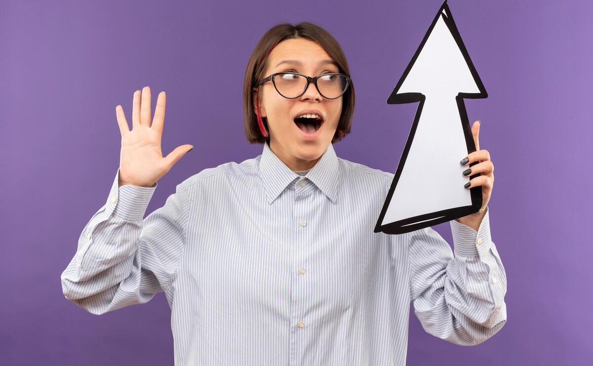 Impressed young call center girl wearing glasses showing empty hand looking at and holding arrow mark which is pointing up isolated on purple