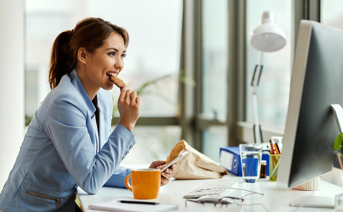 Young businesswoman using cell phone while eating a cookie at her office desk