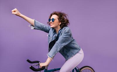 Confident girl in denim jacket riding on bike and waving hand. Indoor photo of inspired young lady in glasses sitting on bicycle.