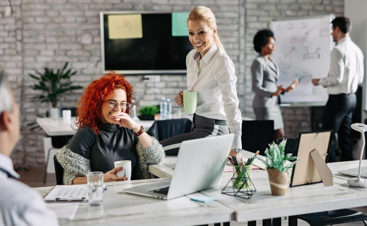 Two businesswomen laughing while talking to their colleague and drinking coffee in modern office There are people in the background