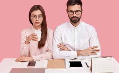 Shot of serious female and female perfectionist, being well organized, wear spectacles, everything on right place at working place, drink coffee, work together on new project, isolated on pink