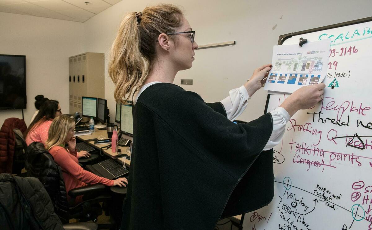 A woman is placing a printed page on an unorganized whiteboard.