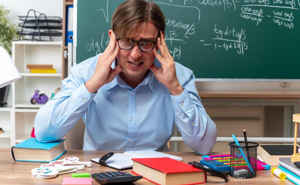 Young male teacher wearing glasses stressed and nervous sitting at school desk with books and notes in front of blackboard in classroom