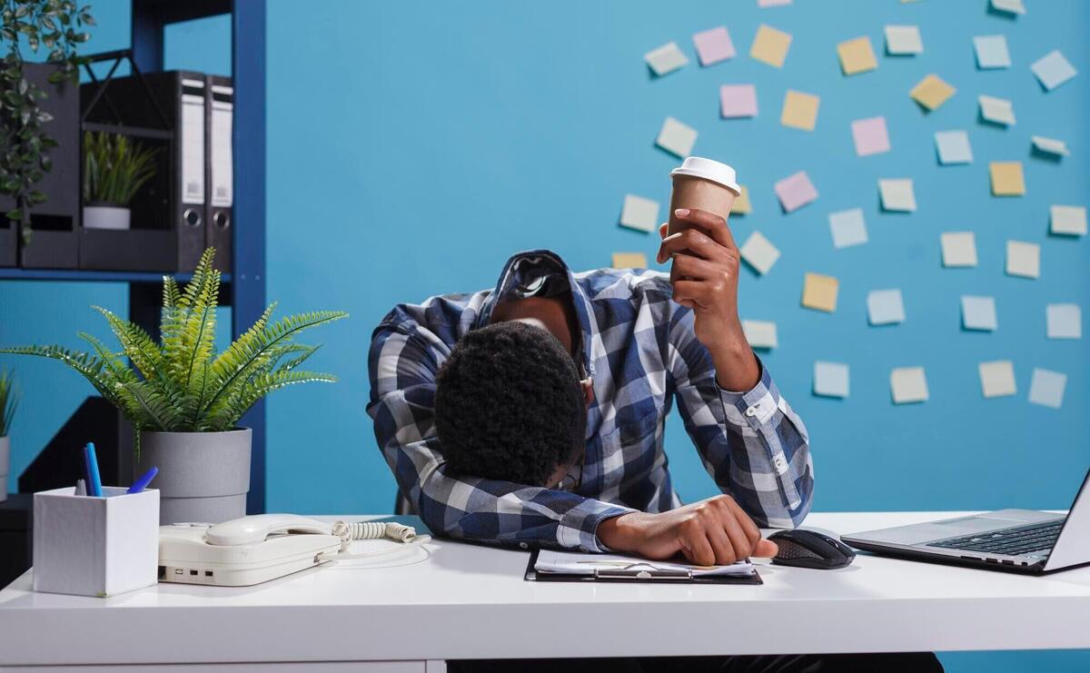 Stressed and fatigued team leader with burnout syndrome sitting in modern office workspace. Tired and exhausted agency office employee falling asleep at desk because of overtime work