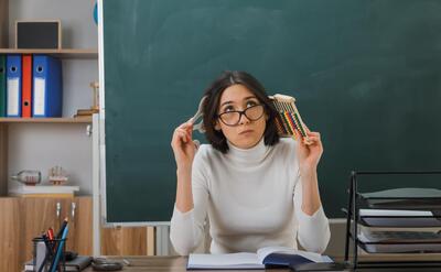 thinking young female teacher wearing glasses holding abacus with number fan sitting at desk with school tools on in classroom