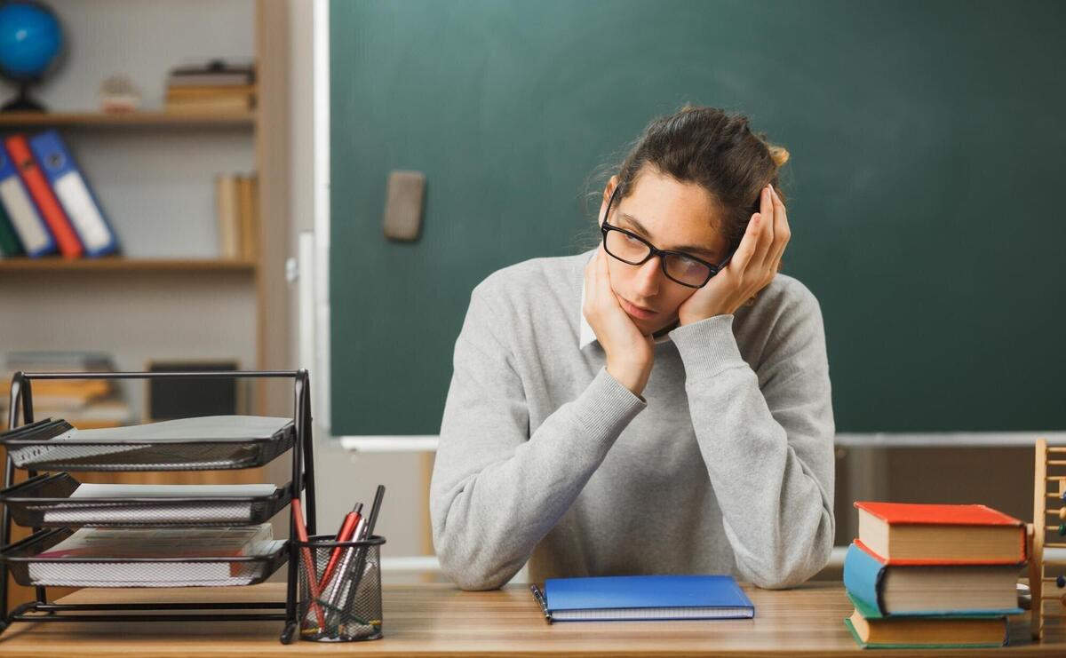 sad grabbed head young male teacher sitting at desk with school tools on in classroom