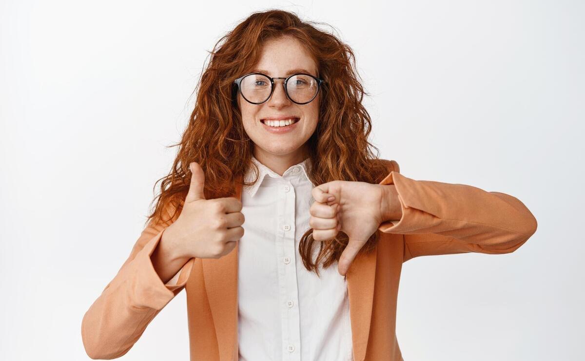 Like or dislike smiling saleswoman with red curly hair showing thumbs up and thumbs down advantage or disadvantage gesture standing in suit and glasses over white background