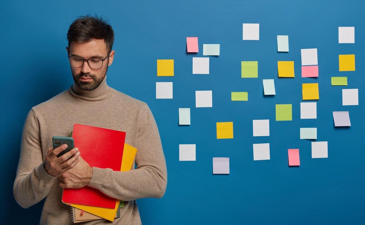 bearded man uses mobile phone for online conversation, holds textbook, wears spectacles, dressed in glasses and brown sweater, looks for information, colorful notes behind on wall