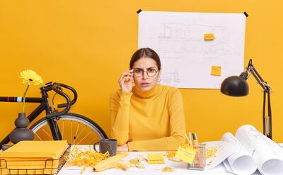 Horizontal shot of brunette woman looks attentively, keeps hand on spectacles tries to see something into distance poses at messy desktop works on drawing for architectural project.